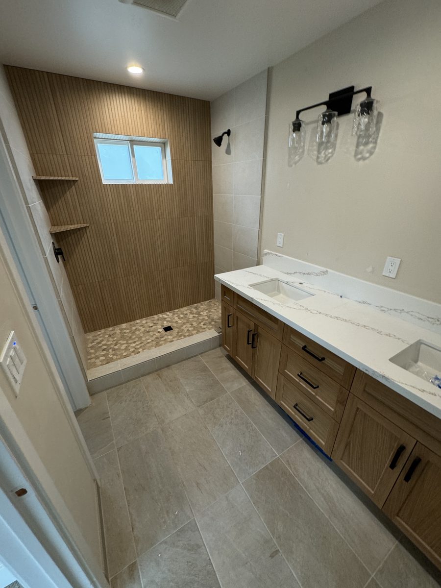 A bathroom with a large gray tile floor, a wood-look tile shower wall and a pebble shower floor beside a double vanity.