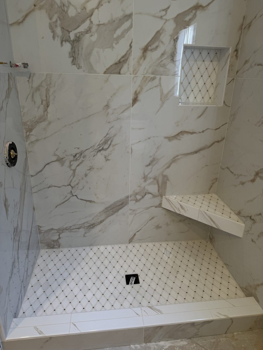 A shower in large marble-look tile with a corner bench, a diamond-pattern niche and a matching mosaic floor.