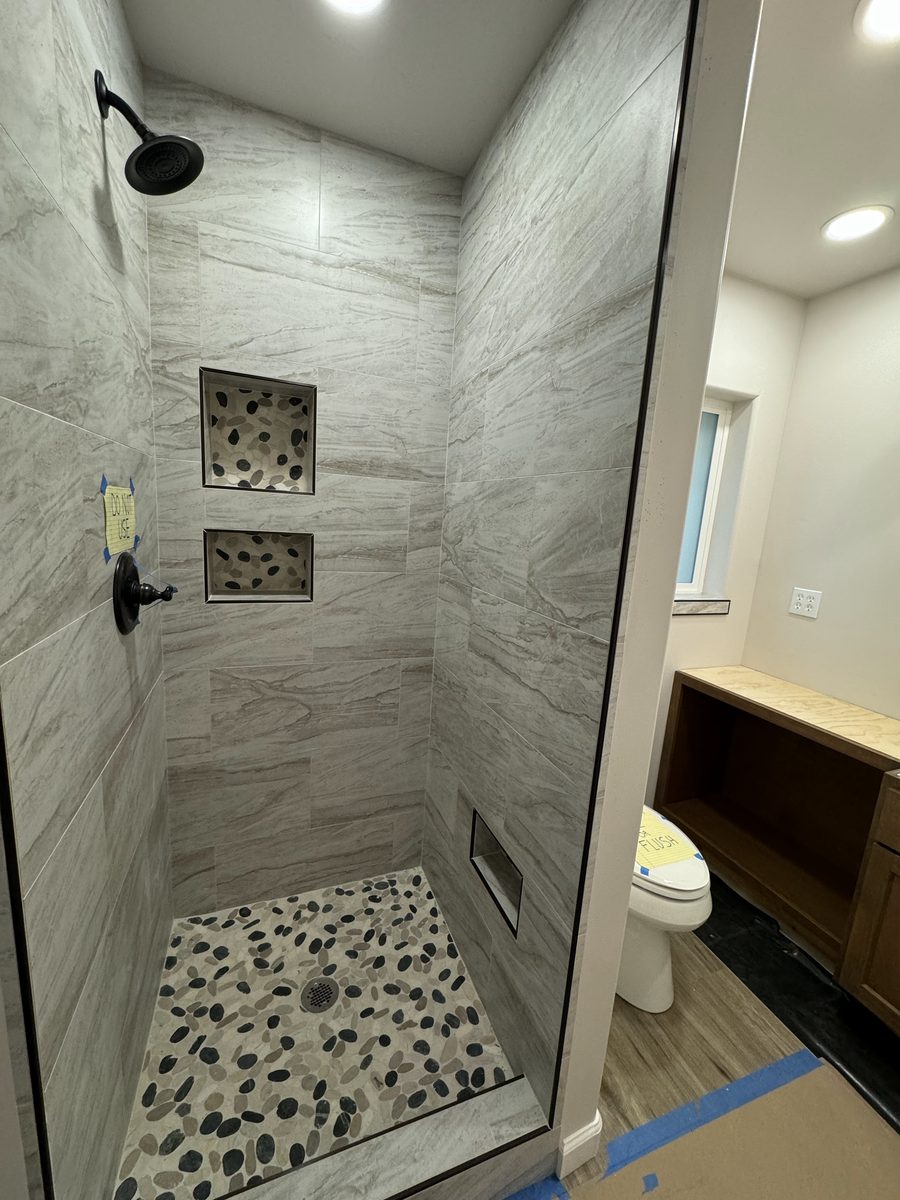 A shower in gray stone-look tile with two pebble-lined niches and a black and gray pebble floor.
