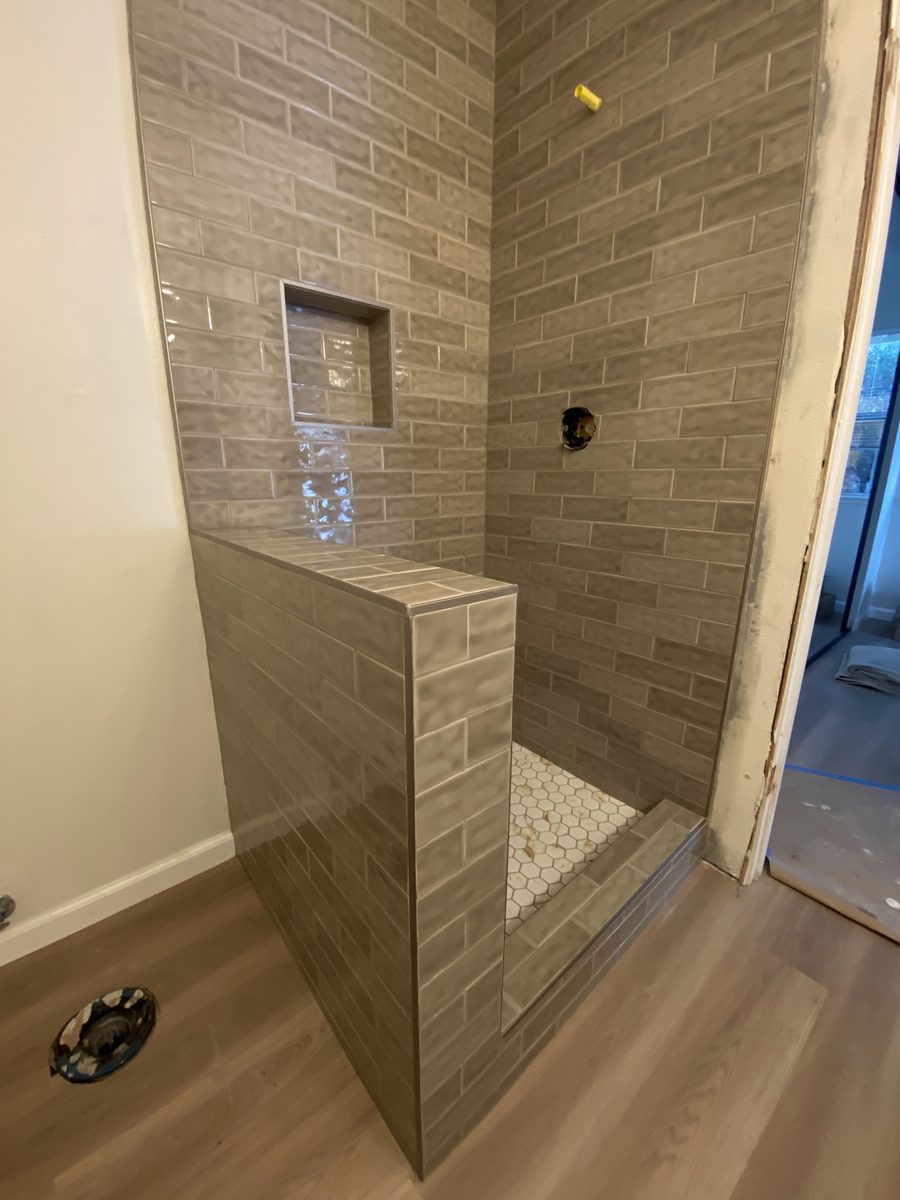 A shower in long taupe plank tile with a half wall, a recessed niche and a mosaic floor.